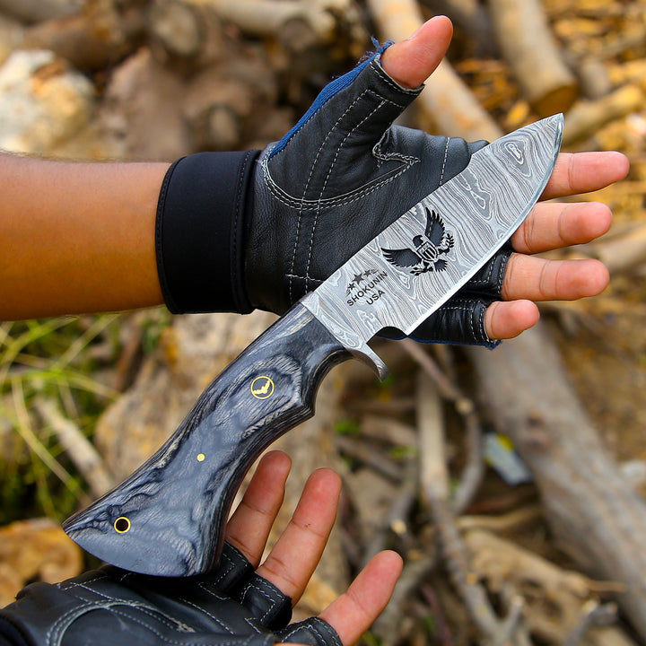 Hand holding a knife with a detailed blade and engraved design, wearing a black glove against a natural background.